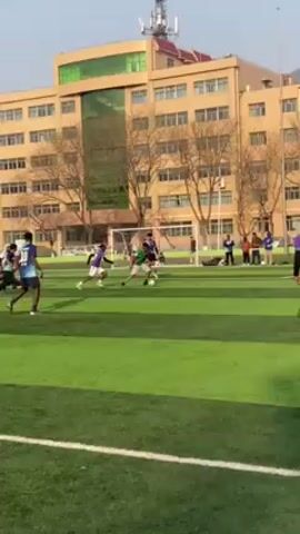 The video shows a group of men playing soccer on a green turf field with a large building in the background. The players are wearing different colored jerseys, including purple, green, and blue. Some players are running while others are kicking the ball. One player is on the ground. The sky is clear and blue.