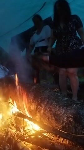 The video shows a group of people gathered around a campfire under a tarp. The fire is burning brightly, with sparks flying. Two individuals, a male and a female, are visible dancing in the background. The female is wearing a patterned dress, and the male is in a white shirt. Several other figures are indistinctly seen in the dim light. The environment appears to be outdoors, possibly a campsite, with foliage visible in the background.