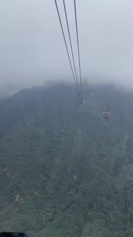 A cable car is ascending a steep, lush green mountain shrouded in fog. The view is from inside another cable car, looking out at the mountain and the cableway. Several other cable cars can be seen in the distance, ascending the mountain. The weather appears overcast and misty. The lower portion of the frame shows the edge of what appears to be a phone screen.