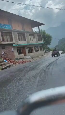 The video captures a scenic view of a mountainous region, possibly during monsoon season. A wet, asphalt road winds through a valley with steep, green-covered mountains on the right. A multi-story building, appearing to be a guesthouse or hotel, is situated on the left side of the road, close to a stream or flooded area. In the distance, a person is riding an ATV on the road. A black dog is walking on the road, seemingly interacting with the ATV or its rider. The sky is overcast with clouds, suggesting potential rainfall or recent rain. The overall atmosphere is one of natural beauty and rugged terrain, with signs of human activity in the form of the building and vehicles.