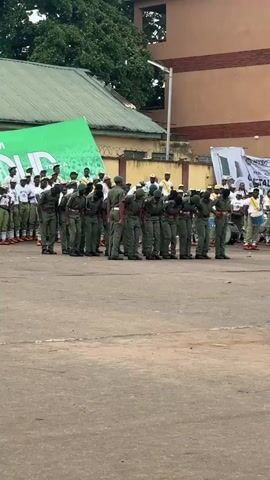 A group of people wearing matching green uniforms and hats stand at attention in a courtyard or paved area. They are bowing their heads in unison. Behind them, a large green banner with white lettering is visible, along with other banners and some buildings. Several other individuals in various attire are also present in the background. The overall scene appears to be an organized event or ceremony.