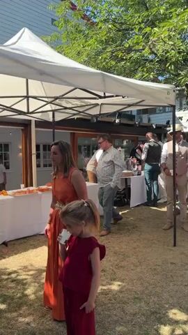 The video captures an outdoor gathering, likely a party or wedding reception. A white canopy tent is set up, providing shade. Several people are visible, dressed in semi-formal and casual attire. Some individuals are standing and talking, while others appear to be interacting near tables laden with food and drinks. A child in a red dress is in the foreground. In the background, there's a building with large windows, lush green trees, and a bright blue sky. The ground is covered in grass and some scattered leaves. A ornate, multi-branched candelabra with lit white candles is prominent on a table in the latter part of the video. The atmosphere appears relaxed and celebratory.