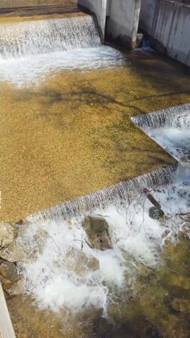 A high-angle shot shows water cascading over two tiered concrete structures into a small stream. The water flows clear over the top tier, creating a foamy white surface, then pours over the edge. The mid-level area is relatively shallow, with a visible rocky bottom under clear water. The water then drops over another concrete tier, churning around several large rocks and a fallen branch. The sun casts shadows on the concrete surface. A concrete structure is visible on the right side of the video, and a small footbridge is visible in the far background.