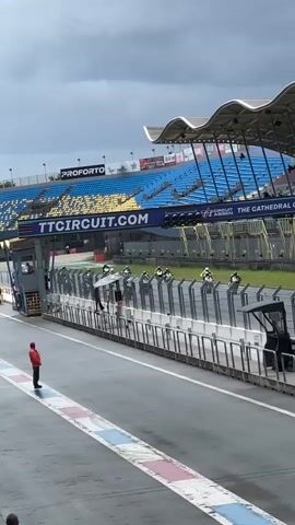 A motorcyclist in a red jacket stands on the left side of a wet asphalt race track. In the background, a row of motorcycles with riders wearing helmets and green and white racing suits are lined up on the track, behind a white barrier. A large grandstand with blue and yellow seats is visible behind the barrier. The sky is overcast. A large banner above the barrier reads "TTCCIRCUIT.COM THE CATHEDRAL OF SPEED".