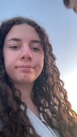 A woman with long curly brown hair is recording a video of herself and a man on a beach. She is wearing a white top. The man beside her has short dark hair and is wearing a dark-colored jacket with a white Nike logo. The sky is light blue and the beach appears to be sandy. The sun is setting. The woman makes a pouting face at the camera. The man looks off to the side.