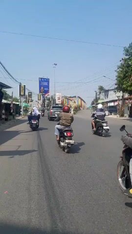 This video depicts a daytime scene on a road, likely in a tropical or subtropical region, given the clear blue sky and lush green trees visible. The road is paved and appears to be a multi-lane highway. Several motorcycles are in motion, with riders wearing casual clothing and some headscarves. One motorcycle in the foreground has two riders, one driving and one passenger. Traffic appears moderate, with other motorcycles and a dark-colored car also on the road. On either side of the road, there are buildings and stalls, suggesting a populated area, possibly a town or city. Billboards are visible along the roadside, one of which is quite large and features a graphic and text. Overhead, power lines are strung between poles, indicating the presence of infrastructure. In the distance, a bridge is visible, adorned with colorful banners or flags. The overall impression is of everyday life and movement in a developing urban or suburban environment.