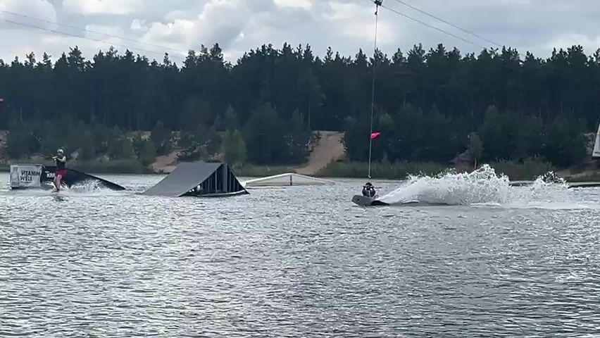 The video shows a wakeboarding park on a lake. Two individuals are wakeboarding. One is on the left side of the screen, wearing a red shirt and black shorts, and is being pulled by a cable. They are near a ramp-like structure. Another wakeboarder is in the center of the screen, wearing a blue shirt and khaki shorts, also being pulled by a cable. They are producing a large spray of water as they ride. Further back in the lake, there are inflatable water structures, including a large yellow and blue slide. The background consists of a dense forest and a sandy embankment. The sky is cloudy. A young girl is visible in the foreground of the last few frames, looking towards the camera.