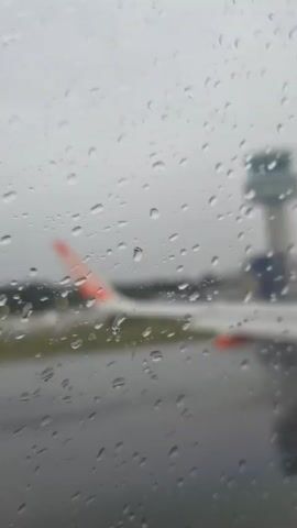 The view from inside an airplane looking out a window shows a plane's wing, a control tower, and rain. Water droplets are on the window. The plane is on the tarmac. An announcement can be heard.