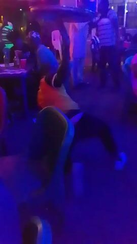 The video shows a room with people sitting and standing under blue lighting with a decorative ceiling. A man in a yellow vest and dark pants stands in the foreground, briefly raising an object over his head before placing it down and walking a few steps. Other people are seen sitting at tables and standing in the background, some wearing casual clothing. The room appears to be a bar or a club due to the lighting and the presence of tables with drinks. There is noticeable noise in the background, possibly music or conversation.