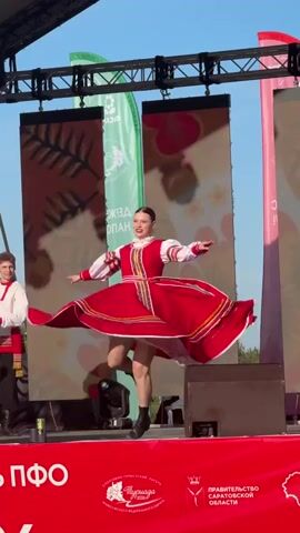 A young woman performs a Russian folk dance on an outdoor stage. She wears a red and white dress, black boots, and her hair is pulled back in a bun. She smiles and executes spins and jumps. A young man in traditional dress stands to her left. The stage is red and features logos and text in white. Behind the dancers are three large screens, flanked by green, white, and pink banners. The sky is blue.