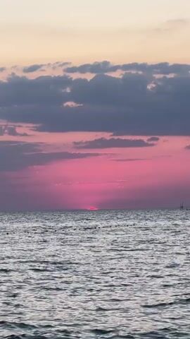 This media file depicts a serene ocean sunset scene. The sky is a vibrant spectacle of pink and purple hues, with streaks of deeper pink emanating from the setting sun, which is partially obscured by the horizon. Dark gray clouds are scattered across the upper portion of the sky, adding depth and contrast to the colorful canvas. Below the sky, the vast expanse of the ocean stretches out, its surface rippled by gentle waves that reflect the colors of the sunset. The overall impression is one of tranquility and natural beauty.

The time of day is clearly sunset, as indicated by the low position of the sun and the characteristic colors in the sky. The weather appears to be calm, with no signs of strong winds or harsh conditions. The setting is coastal, likely a beach or a viewpoint overlooking the sea. The presence of a distant, thin pole-like structure on the right side of the frame might suggest a buoy, a marker, or a part of a distant pier or structure.

The emotions evoked by this scene are likely peacefulness, awe, and a sense of wonder at the beauty of nature. There are no people or objects in the foreground or mid-ground, allowing the viewer to focus entirely on the interplay of light, sky, and sea. There is no visible text in the media file.