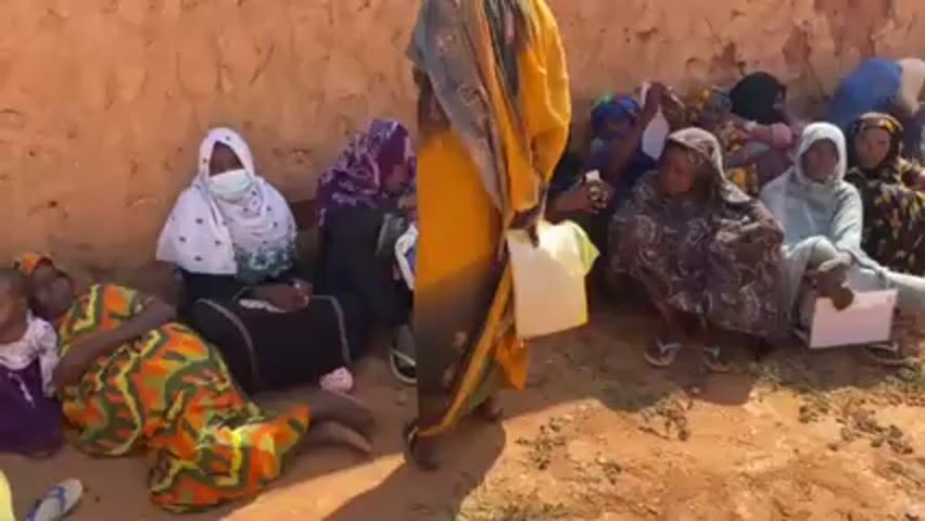 A group of around a dozen women are sitting on the ground in front of a low wall, possibly a dirt wall, outside in bright sunlight. Most of the women are wearing head coverings of different colors and patterns. One woman near the front is wearing a white face mask. Many of the women are holding pieces of white paper in their laps. A woman wearing a yellow and dark gray patterned robe stands in front of them, holding a white plastic container. The ground is mostly dirt.