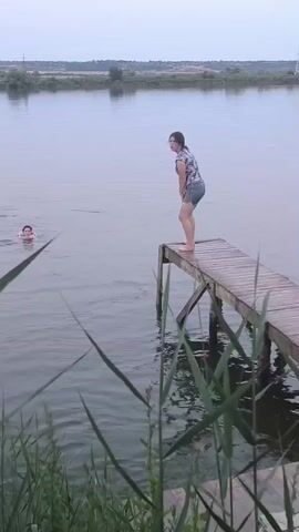 A person stands at the end of a wooden pier, facing away from the camera and looking out at a body of water. Another person is in the water to the left of the frame, partially submerged. The person on the pier bends forward and jumps into the water. There is a splash as the person enters the water. The pier extends from the right side of the frame towards the center. Reeds and tall grass are in the foreground. The sky is overcast.