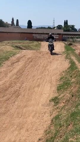 A motorcyclist wearing black riding gear and a helmet rides along a dirt track on a sunny day. The motorcycle has a white front fender and a black seat. A man sits on the side of the track with a camera. The track has small hills and is surrounded by grass and some trees. In the background, there is a building and a few other people. The motorcyclist rides past the camera and then turns away, riding up the hill, past a motorcycle parked on the side, and two people looking at the rider.