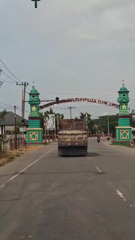 A truck and a motorcycle are driving on a road. There is a large archway over the road with text on it. The archway has two towers on either side, painted green and red. There are palm trees and other foliage in the background, as well as utility poles and wires. The sky is overcast.