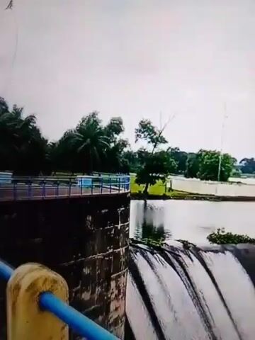 The video shows a dam with water flowing over it. There are trees and a cloudy sky in the background.