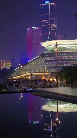 The video captures a night scene of a city with brightly lit skyscrapers and a modern-looking building with a curved roof. A calm body of water in the foreground reflects the lights of the buildings and the sky. Several people are walking along a paved pathway beside the water. Two individuals, a man and a woman, are walking towards the right of the frame. The man wears a light-colored shirt and dark pants, while the woman has on a dark outfit. Another person is further to the right, facing away from the camera. In the background, the architecture is illuminated with colorful lights, including a tall, slender skyscraper with vertical lighting and a shorter, wider building with a patterned facade. The overall atmosphere is one of an urban nightscape with pedestrian activity.