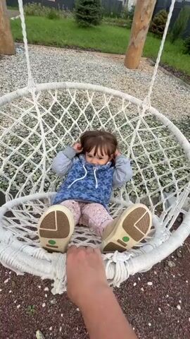 The video begins with a close-up of a young child sitting in a white macrame swing chair. The child is wearing a blue hooded sweatshirt with white stars and pink pants. The child is looking at the camera and holding their hands near their head. The camera then pans out to show the child swinging gently. The background is a yard with gravel, some rocks, and wooden posts supporting the swing. In the distance, there are houses and trees. The video ends with a view of the child still in the swing.