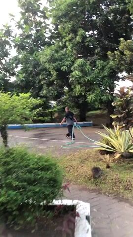The video shows a man with dark hair and a dark shirt standing in an outdoor area with trees and vegetation. He is holding a green hose. The ground appears to be paved with interlocking bricks and some areas of grass and dirt. There is a blue and white structure in the background.