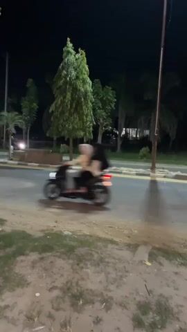 The video captures a nighttime scene on a paved road in Kebuntanahterban, Indonesia. The setting appears to be a roadside with trees and palm trees lining one side, and a grassy, sandy area on the other. A prominent street lamp casts a shadow on the road, indicating artificial lighting.

The primary activity involves several motorcycles traveling along the road. In the early frames, a motorcycle with its headlight on is visible moving from left to right. Later, other motorcycles, some with riders and possibly passengers, are seen in motion. The road has painted lane dividers. There are also some low concrete barriers or planters along the side of the road.

The time of day is clearly night, with a dark sky and illuminated by artificial lights from the street lamp and the vehicles. The weather appears clear, as there's no indication of rain or extreme conditions. The overall mood is one of transient movement on a quiet road at night. There is no visible text in the video.