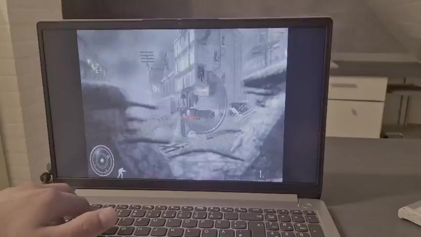 A person's left hand is visible resting on the keyboard of an open laptop. The laptop screen displays a video game scene viewed through a sniper scope. The in-game environment appears to be an outdoor urban setting with brick buildings. A character is visible in the crosshairs, and the player is actively engaged in gameplay, indicated by the hand's position on the keyboard. The overall scene suggests a person playing a first-person shooter or similar genre video game.