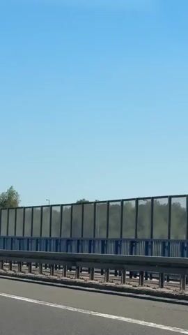 The video shows a view from a vehicle traveling on a highway. On the right side of the highway, there is a sound barrier with a blue metal frame and transparent panels. Beyond the sound barrier, there are trees and a grassy hillside. A tall electricity pylon is visible in the distance on the left side of the frame. The sky is clear and blue. The video appears to be filmed during the daytime, likely in the summer due to the lush greenery.