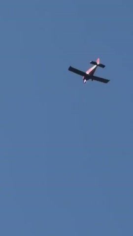 A small, red and white airplane flies across a clear blue sky. The plane is seen from below and appears to be in level flight. The sun glints off its wings and fuselage.