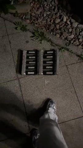 The video shows a pair of feet wearing silver shoes and white pants standing on a black doormat. The doormat is placed on a paved surface next to a pile of rocks and some greenery. The perspective is from above, looking down at the feet and the doormat. The scene appears to be outdoors and dimly lit, possibly at night. The person's feet shift slightly on the doormat.