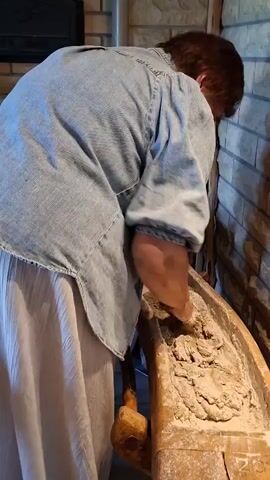 A person wearing a blue denim shirt and a light-colored skirt kneads dough in a wooden trough. The trough is old and appears well-used, with flour dusting its surface. The person's hands are actively working the dough, mixing it with the flour. The background suggests an indoor setting with a stone or brick wall and possibly a fireplace. The lighting is dim, creating shadows that highlight the textures of the dough and the surroundings.