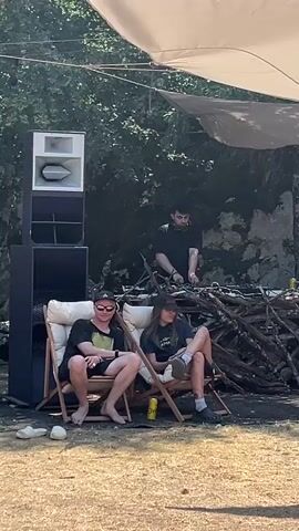 A DJ in a black t-shirt stands behind a DJ setup, with a large speaker system to the left and a pile of branches and rocks to the right. Two people, one wearing a black t-shirt and sunglasses and the other a black t-shirt and a baseball cap, sit in chairs facing the DJ. Both individuals are barefoot. Several yellow cans are visible on the ground near the chairs. The scene appears to be outdoors, under a large canopy, with trees and foliage in the background.