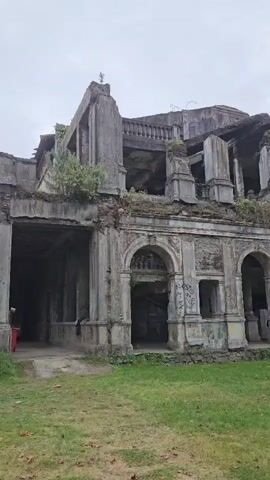 The camera provides a wide, low-angle shot of the deteriorated shell of an old, multi-storied building. The structure is constructed of grey stone and concrete and features arched entryways on the ground floor, with columns supporting the upper levels. The roof is partially collapsed, and the interior is exposed. Vegetation grows on parts of the structure, and graffiti is visible on some walls. A grassy lawn stretches out in front of the building, with a few scattered rocks or debris. The sky is overcast and grey.