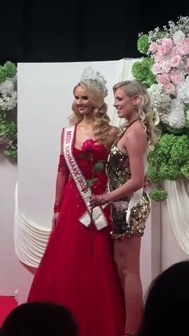 Two women are standing on a red carpet, posing for a picture. The woman on the left is wearing a large silver crown, a long, red, formal gown with a single sleeve and a "Miss Denmark 2023" sash. She holds a red rose. The woman on the right is wearing a short, gold sequined dress and a "Miss Denmark" sash. She is smiling and has her arm around the other woman. Behind them is a white backdrop decorated with pink and white roses and green foliage. The video is slightly shaky.