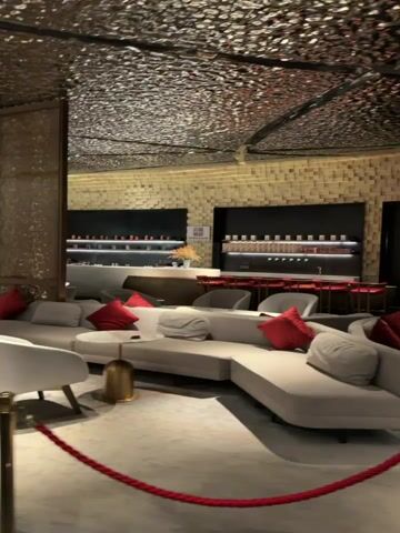 The video pans across a well-lit, modern lounge area with luxurious seating. The walls are a textured beige, and the ceiling has a metallic, hammered finish. Several light gray, curved sofas with red throw pillows are arranged throughout the space, accompanied by white chairs and small, round marble-topped tables with gold bases. In the background, a bar area is visible with a dark countertop, shelves stocked with bottles, and red stools. Red velvet ropes on gold stanchions mark off an area in the foreground. The overall impression is one of a sophisticated and comfortable seating area, possibly in a hotel lobby, upscale bar, or lounge.