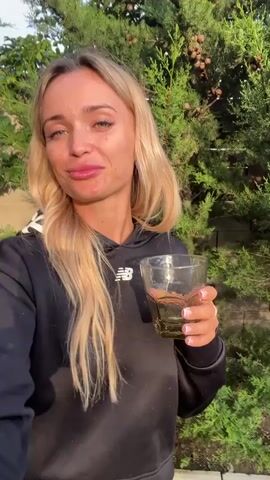 A young Caucasian woman with blonde hair and blue eyes, wearing a black hoodie and black sweatpants, holds a glass with a dark liquid in her right hand. She is standing outdoors in front of a large green conifer tree. The ground around her is covered with small gray stones and some green grass. The lighting suggests it is late afternoon or early evening. The woman is looking at the camera and appears to be speaking.