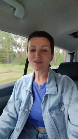 A woman with short, dark hair, wearing a blue t-shirt and a denim jacket, is sitting in the passenger seat of a car, looking at the camera and smiling. The car is in motion, with trees and foliage visible through the window. The woman is gesturing with her hands as she speaks.