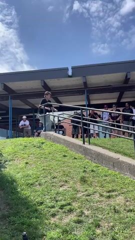 The video shows a group of young people outdoors on a sunny day. In the foreground, a person is riding a scooter down a grassy slope towards a concrete path. They are wearing a black t-shirt, blue jeans, and sneakers. The scooter is metallic with a black handlebar and wheels. The person appears to be performing a trick, sliding on a metal railing that runs alongside a set of stairs. In the background, a group of spectators are gathered, some sitting on the grass, others standing. They are watching the scooter rider. Behind the spectators is a building with a dark overhang and a sign that reads "SNACK BAR". There is graffiti art on the wall of the building. The sky is blue with some white clouds. The overall scene suggests a casual outdoor gathering or a scooter event.