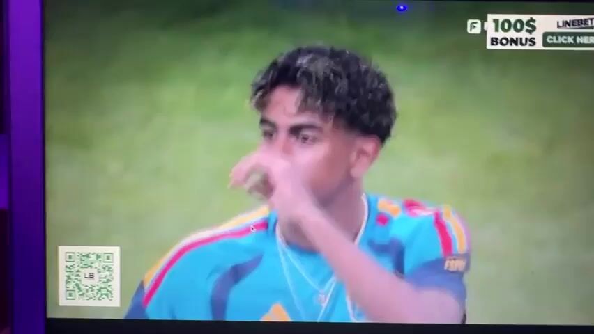 The video shows a young man with curly, highlighted hair, wearing a teal and red striped soccer jersey. He is on a soccer field, and in the background, a large stadium crowd is visible. There is a QR code in the lower left corner of the screen, and a banner with the text "EARN BIG CLICK HERE!" in the upper right corner. The young man appears to be speaking, but his words are not audible. The overall scene is a broadcast of a soccer match.