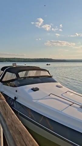 The video begins with a close-up of a white and dark-hulled motorboat docked next to a wooden railing. The boat has a black canopy over the cockpit. The camera then pans right, showing the boat moving away from the dock. The water is calm with gentle ripples. In the background, across the body of water, is a distant shoreline with trees and some industrial structures, including cranes. The sky is blue with scattered white clouds, and the sun is visible, casting a bright reflection on the water. There are a few other small boats visible in the distance. The video captures a serene scene on the water, possibly during sunset due to the warm light.