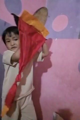 A young child is standing in front of a pink and purple polka-dotted wall. The child is wearing a light-colored t-shirt and pants. In the first few seconds, the child is holding a red and yellow fabric object above their head. They move the object around, and it obscures their face for a moment. The child then lowers the object, revealing their face and a smile. They bring their hands together in front of them. The child appears to be looking directly at the camera.