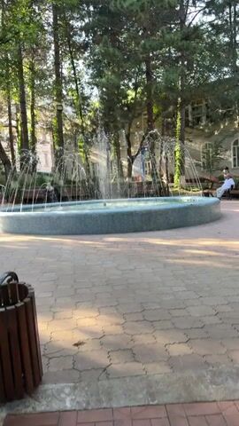 A fountain sprays water in a park-like setting. Trees surround the fountain, and a tiled walkway leads to it. Benches are visible along the perimeter of the fountain, and one person is seated on a bench in the background. The overall atmosphere appears peaceful and serene.