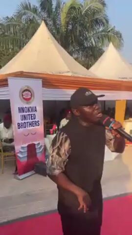 Here is a description of the video:
A man wearing a black cap, patterned shirt, and black vest is speaking into a microphone in front of a red carpet. Behind him, a banner reads "Nnokwa United Brothers". In the background, there are two white tents, and men wearing white outfits and red caps are seated. Palm trees are also visible in the background. The man with the microphone appears to be addressing the group.