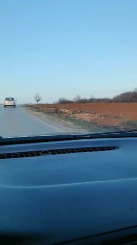 A car is driving on a rural road. The road has a white dividing line and a few cracks. The car is following another white car. On either side of the road are plowed fields and scrubby trees. The sky is clear and light blue. The perspective is from inside the car, looking over the dashboard. The windshield is partially visible, with some streaking.