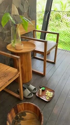 The video shows an outdoor massage space with a wooden floor.  There are two wooden chairs and a small wooden table with a plant on it.  Also on the floor are a pair of slides and a tray with a small bottle and round cut citrus fruits.  A massage table with a patterned cover and a headrest is visible, with a dark bathtub in the background. The massage area is surrounded by a net that offers a view of a lush green environment, including trees, palms, and a rice paddy.