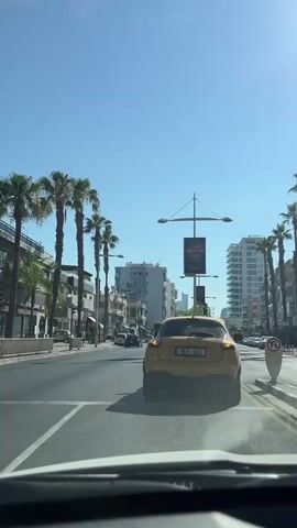 The video shows a street scene from inside a car. Palm trees line the street, with buildings on either side. Several cars are visible, including a yellow car in front of the camera, a white car, and a red car. A sign for "Ford" and "Motion" is visible on one of the buildings. The sky is clear and blue, indicating a sunny day. The video appears to be taken in a city, likely in a warm climate. There are no people clearly visible in the video.