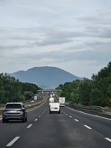 The video shows a view from a car driving on a highway. The highway has multiple lanes of traffic. There are several vehicles on the road, including cars and trucks. The weather appears to be overcast, with a cloudy sky. In the background, there are mountains and trees. The perspective is from the driver's seat, looking forward.