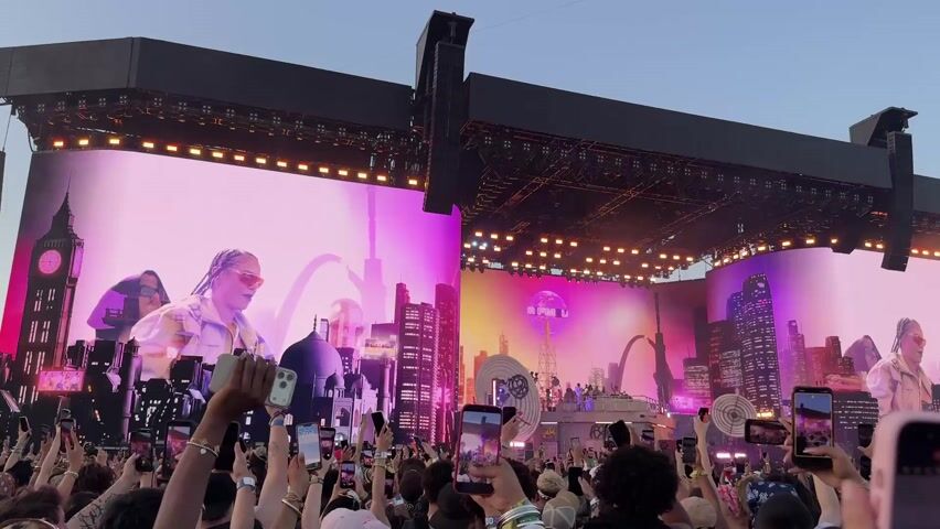 This video captures an outdoor music concert or festival scene from the perspective of the audience. A large crowd of people is visible, mostly from behind, with many holding up their mobile phones to record the event.

The focal point is a massive stage featuring multiple large LED screens displaying dynamic visuals. The screens show a main performer wearing sunglasses and a head covering, along with animated cityscapes that include landmarks like Big Ben, Middle Eastern-style architecture, modern skyscrapers, and a radio tower. On one of the screens, the text "SUPER FM LA" is visible on the radio tower. Below the main screens, small figures of performers can be seen on the actual stage, with one person in glasses appearing to be behind a DJ console or similar equipment.

The stage is equipped with extensive lighting fixtures and large speaker arrays hanging from the truss structure. The sky above is a clear light blue, suggesting late afternoon or early evening. The overall atmosphere appears energetic, with the crowd engaged in the performance.