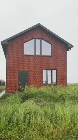 The video shows a two-story brick house with black door and window frames, located in an overgrown field under an overcast sky. The camera pans from the house to a sign in the field that reads "DOM NA PRODAZHU" (House For Sale) along with a QR code and phone number. Tall buildings can be seen in the distance behind the sign and a telecommunications tower extends high into the cloudy sky.