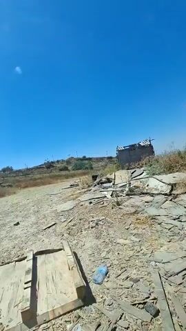 The video shows a dry, outdoor environment under a clear blue sky. The ground is covered in gravel and debris, with some scattered wooden planks and a plastic bottle visible in the foreground. In the mid-ground, there is a slightly elevated area with dry vegetation and a dilapidated structure on the right. Beyond this, a hillside is visible with some trees and scattered buildings or structures. Several flags are flying on poles on the hillside, and there appears to be a fence or barrier running across the mid-ground. The overall impression is of a somewhat neglected or desolate rural or semi-urban area.