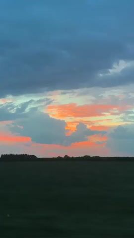 The video shows a view from a moving vehicle of a sunset over a rural landscape. The sky is filled with dramatic clouds, with streaks of orange and pink from the setting sun visible. Below the sky is a dark green field, with a line of trees in the distance. The top of the video frame is partially obscured by the interior of the vehicle.