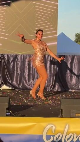 A young woman in a sparkly gold costume with fringe detail is dancing on a stage. She has her hair styled up with a beaded headband and is wearing high-heeled shoes. The stage has confetti scattered on it. Behind her is a dark blue curtain and a large screen displaying a geometric pattern in green and white. There are speakers on either side of the stage. In the foreground, there's a yellow banner with the word "Color" written in script. The lighting suggests it is daytime or dusk.